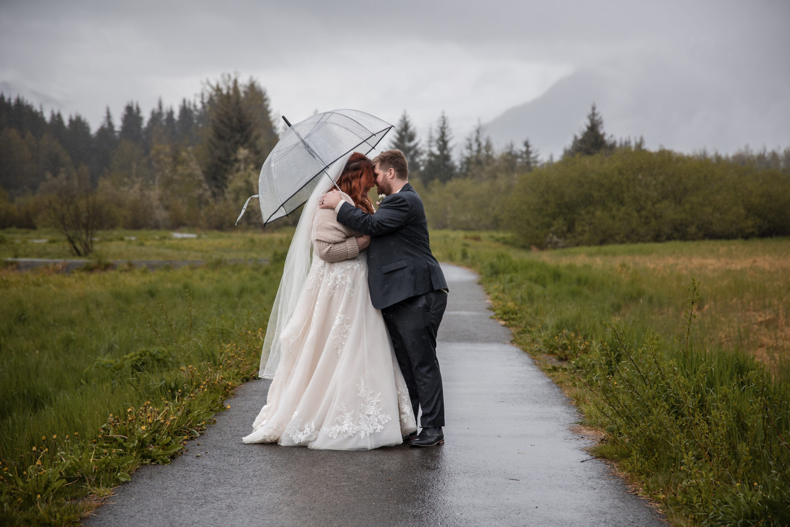 Couple stands on a path on their wedding day under an umbrella kissing during their elopement in Juneau, Alaska