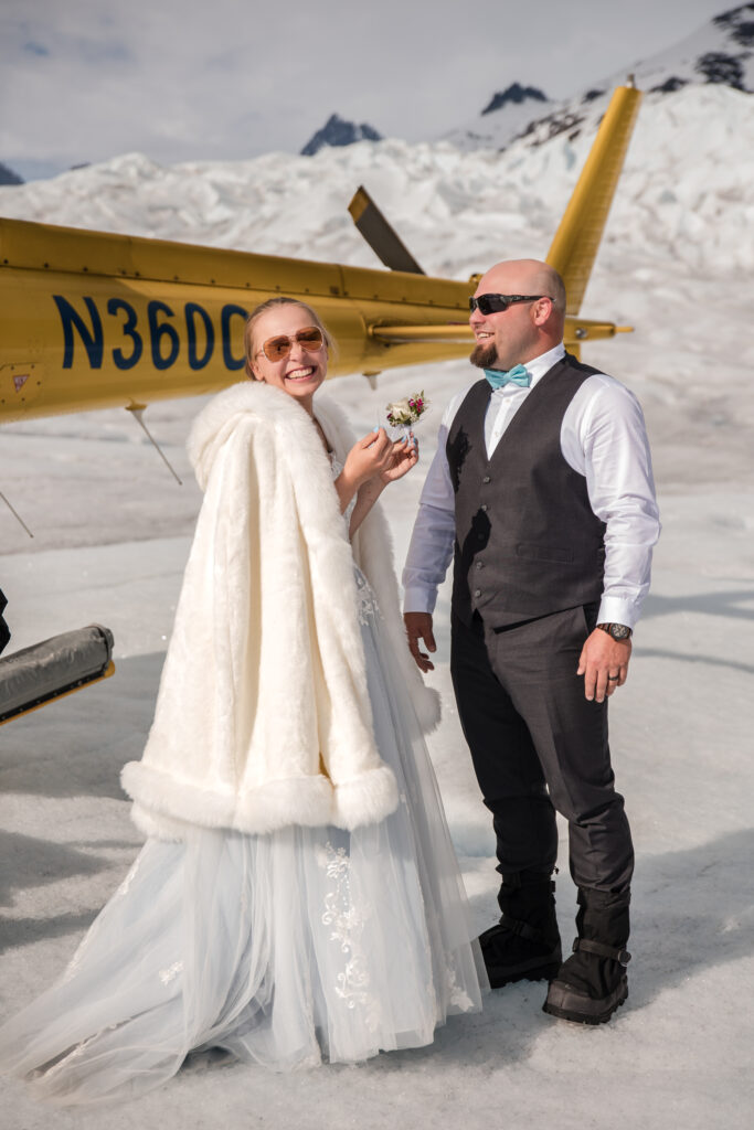 Bride in a white fur cape and groom in a dark vest stand beside a yellow helicopter on a snow-covered glacier.