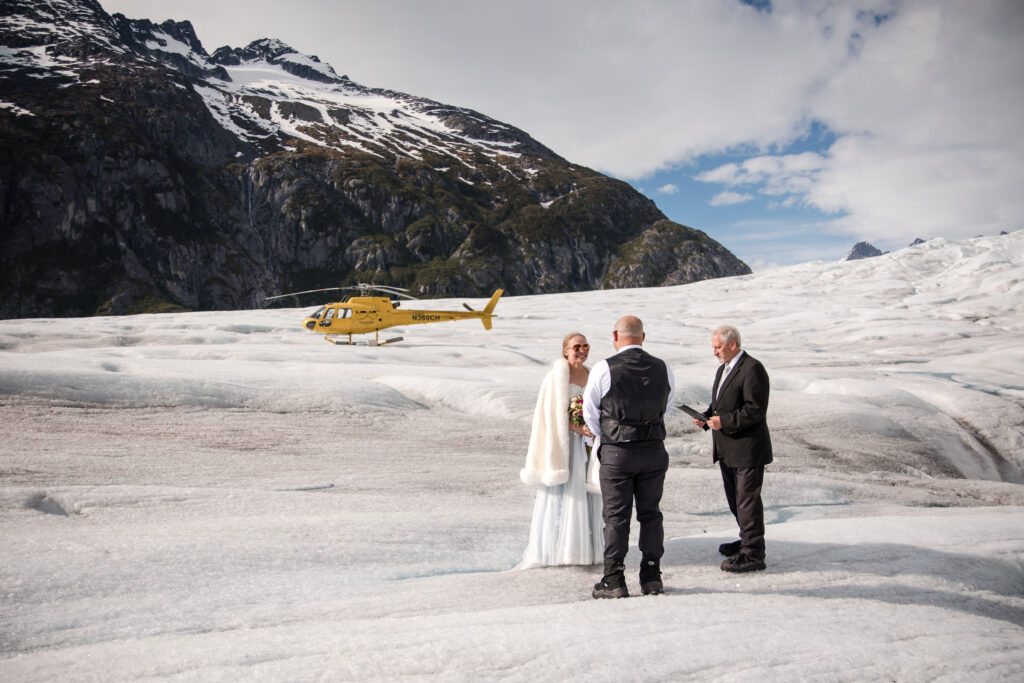 couple exchanging vows on top of glacier in Alaska for elopement with helicopter and mountains in the background