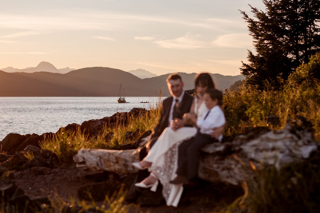 Newlyweds and a child sit on driftwood along an Alaska shoreline at sunset, with mountains across the water.