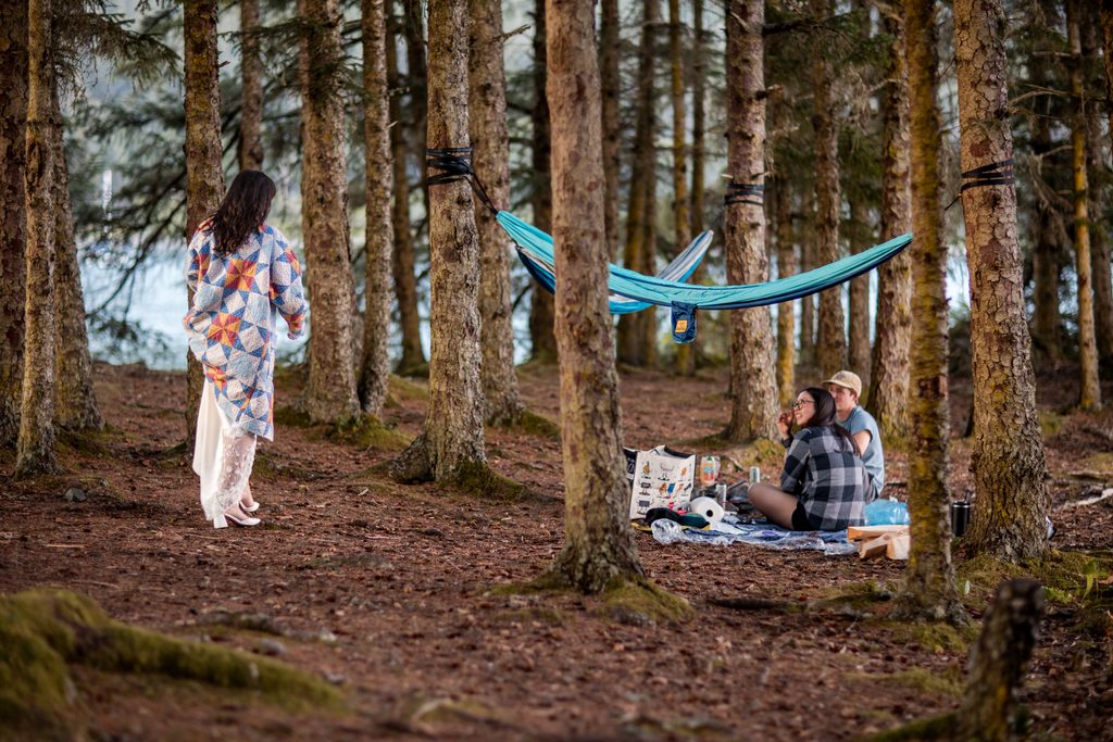 Couple relaxes on a picnic blanket beneath a turquoise hammock as the bride approaches through the forest to ask them to be their witness.