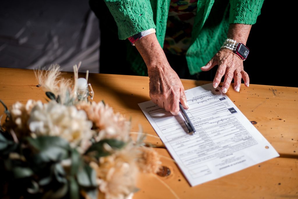 Person points to a section of an Alaska marriage license on a wooden table beside a bridal bouquet.