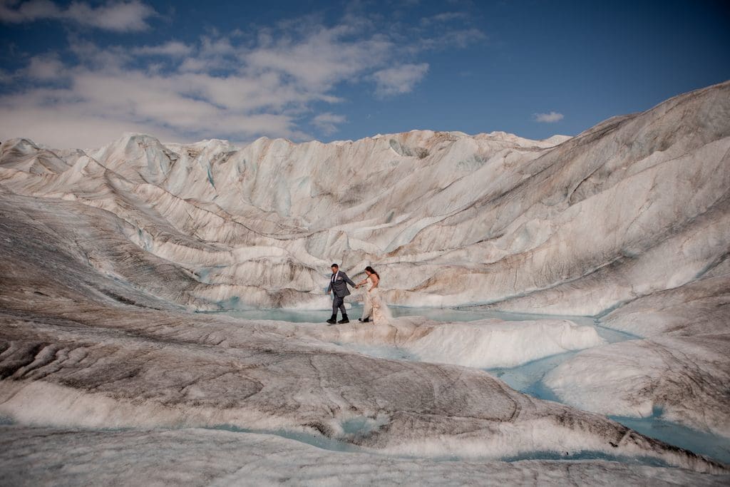 Couple holds hands on a ridge of blue ice surrounded by sweeping glacier walls beneath a partly cloudy sky.