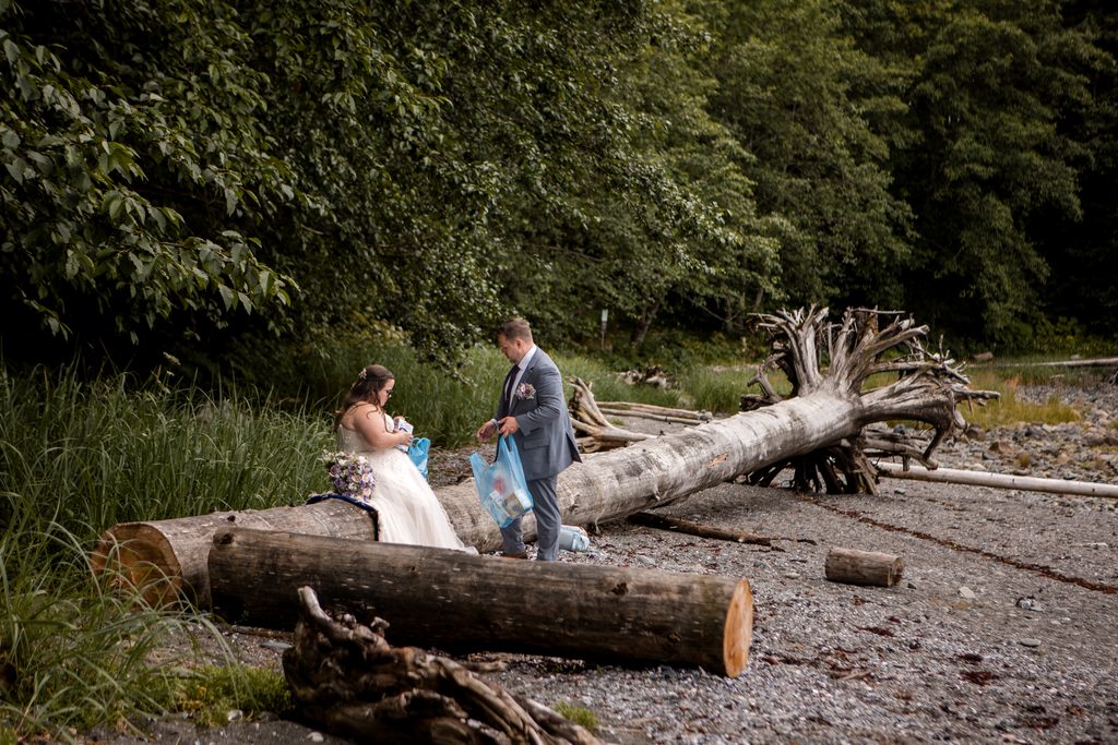 Couple stands beside large driftwood logs on a rocky Alaska beach bordered by lush forest.