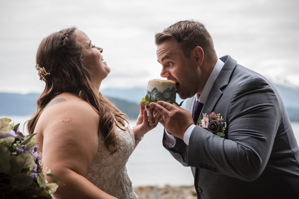 Laughing bride watches her partner take a bite from a small wedding cake beside the water.