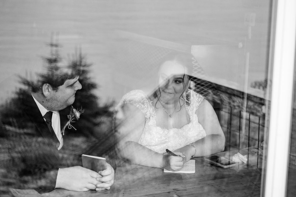 Black-and-white photo through a window of a bride writing in a vow booklet beside her partner, with trees reflected in the glass.