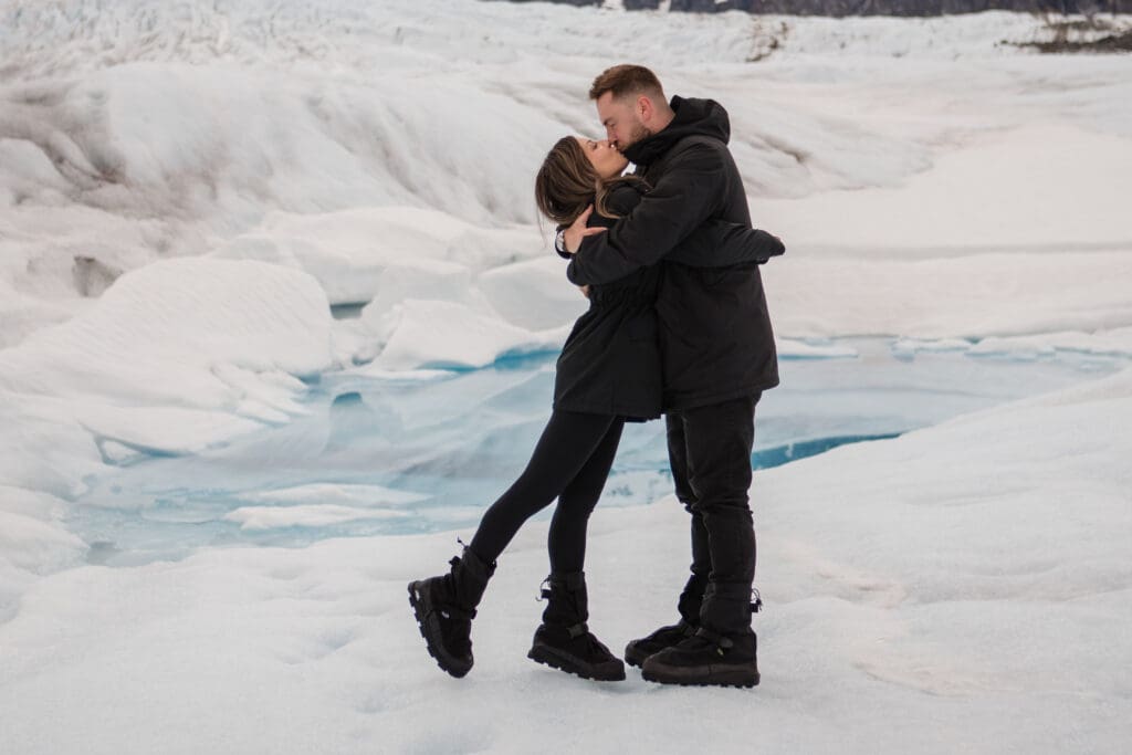 Couple wearing black coats and crampons kisses on glacier ice beside a turquoise meltwater pool.