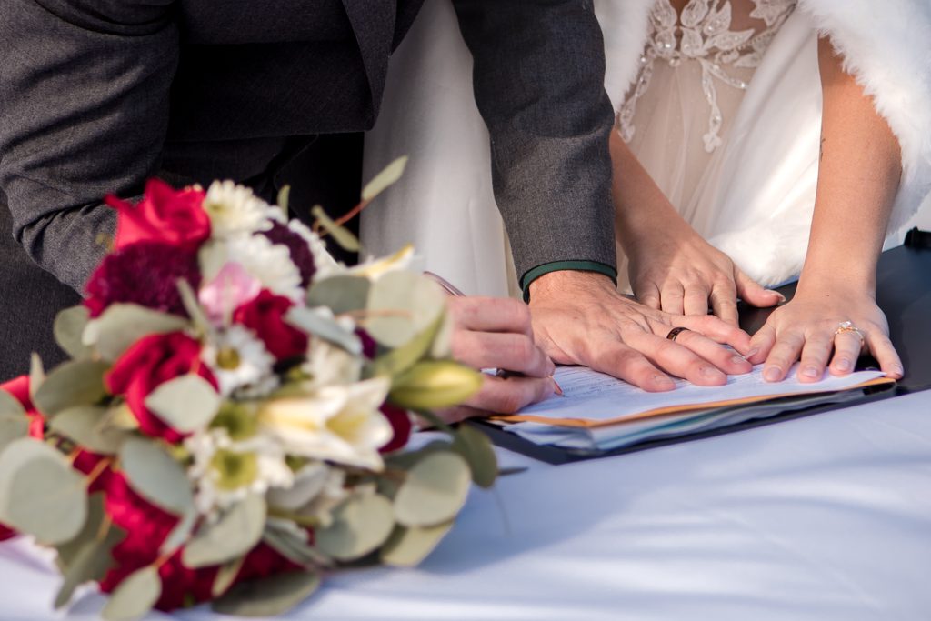 Close-up of a couple signing their Alaska marriage license beside a bouquet of red and white flowers.