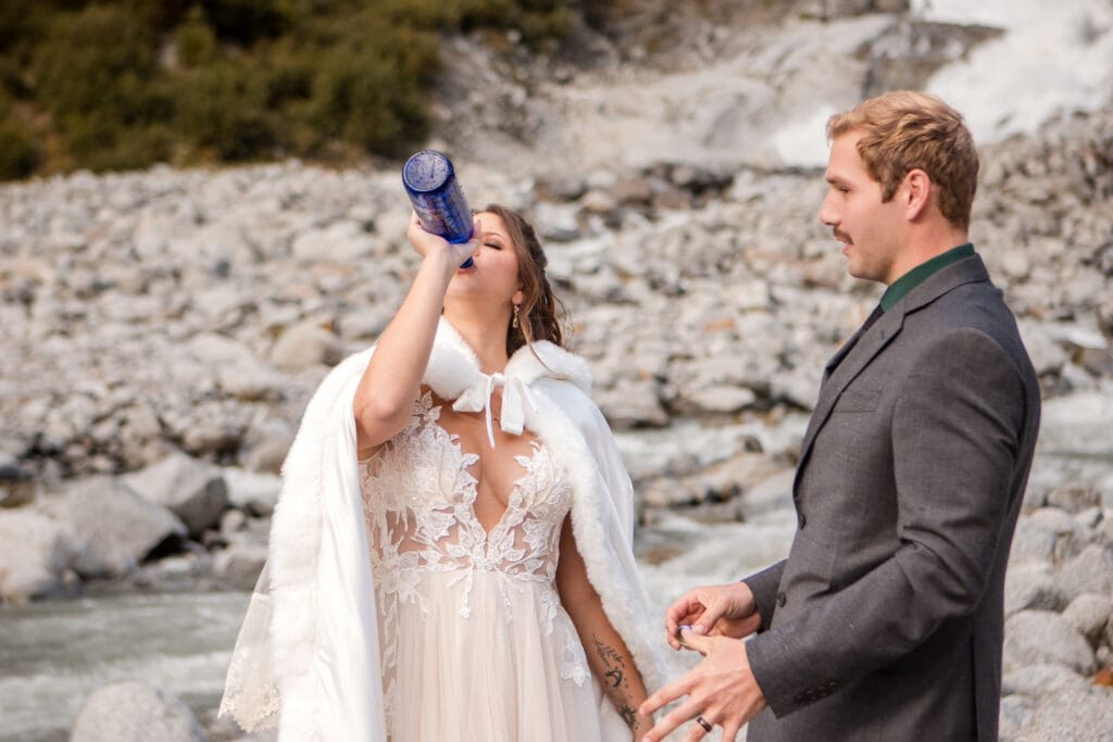 Bride drinks from a blue champagne bottle while holding her groom’s hand beside a rocky glacier stream.