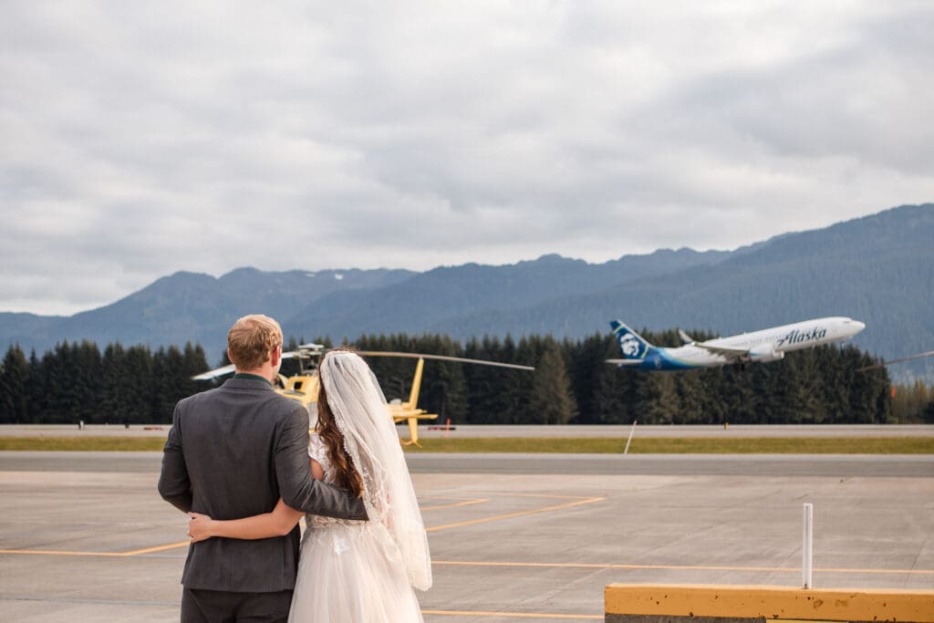 Couple embraces on the airport tarmac before their Juneau helicopter elopement as an airplane takes off behind them.