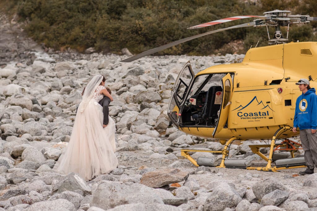 Bride get a piggy back from groom to a yellow Coastal helicopter on rocky terrain during the second stop on their Juneau helicopter elopement.