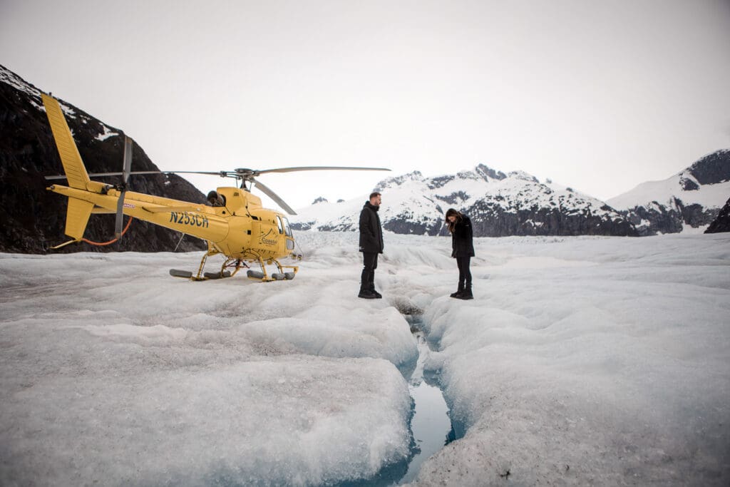 Couple stands beside a blue glacier crevasse and a yellow helicopter during their Juneau helicopter elopement.