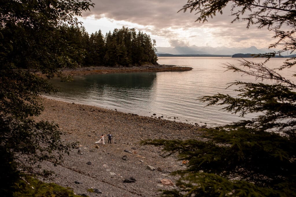Wide view of a couple on a secluded Juneau beach, surrounded by evergreen forest and calm water at sunset.